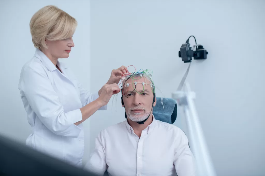 Female-neurologist-adjusting-electrodes-on-gray-haired-male-patient-head-undergoing-electroencephalogram-scaled Female neurologist adjusting electrodes on gray-haired male patient head undergoing electroencephalogram