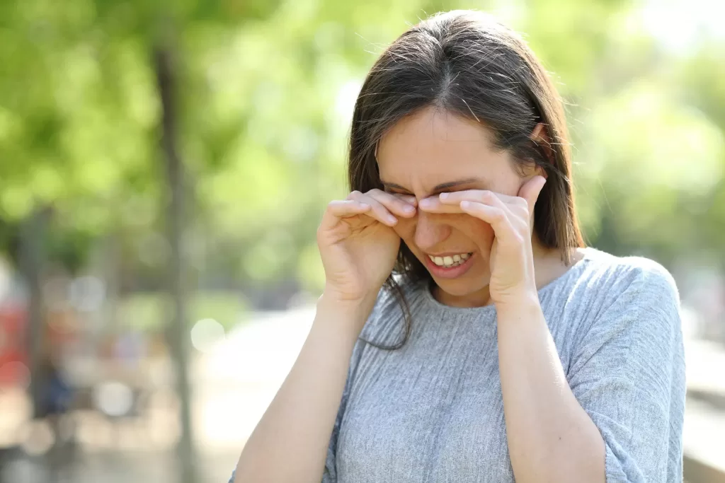 Woman with allergy rubbing her eyes while standing outdoors in a park Woman with allergy rubbing her eyes while standing outdoors in a park