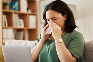 Remote,Work,,Laptop,And,Sick,Woman,On,A,Sofa,With Sick girl wrapped in scarf holding tissue at face and consulting doctor online on video call